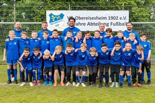 Gruppenfoto von jungen Fußballspielern in blauen Trikots und Trainern vor einem Vereinsbanner auf dem Fußballplatz.