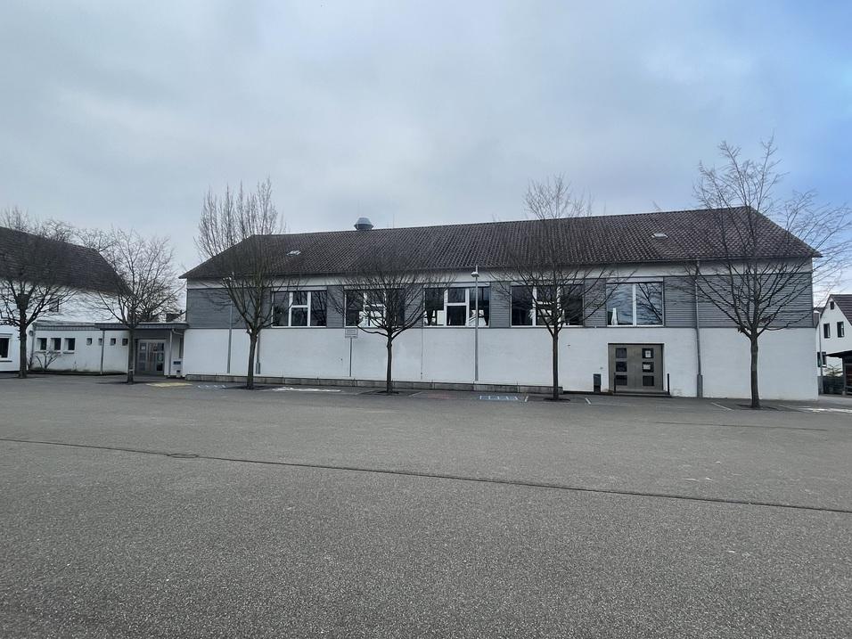 Einfamilienhaus mit grauer Fassade und Fenstern, umgeben von kahlen Bäumen auf einem Parkplatz bei bewölktem Himmel.