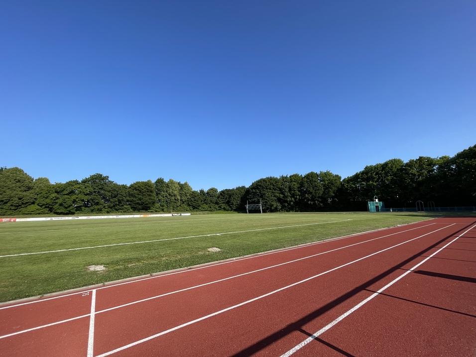 Sportplatz Leere Laufbahn mit rotem Belag und gepflegtem Rasenplatz, umgeben von Bäumen unter klarem, blauem Himmel.