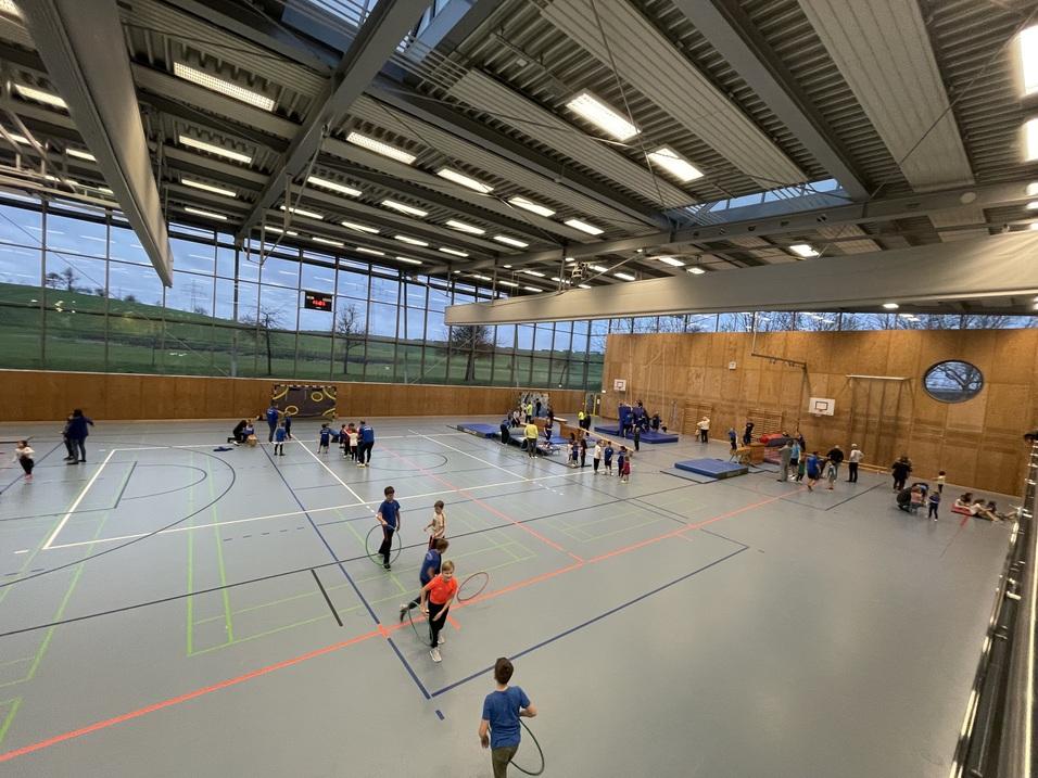 Eberwinhalle Blick in eine Sporthalle mit Kindern, die verschiedene Aktivitäten ausführen, z.B. mit Bällen und Turngeräten.