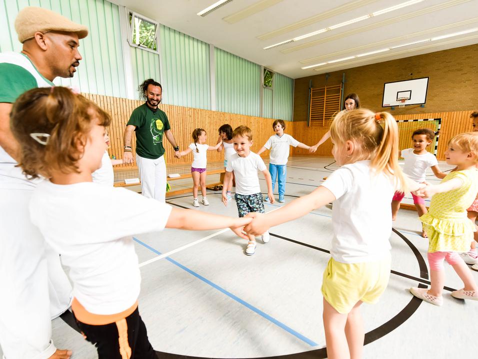 Gruppe von Kindern in T-Shirts und Erwachsenen in einer Turnhalle, die gemeinsam einen Kreis bilden und spielen.